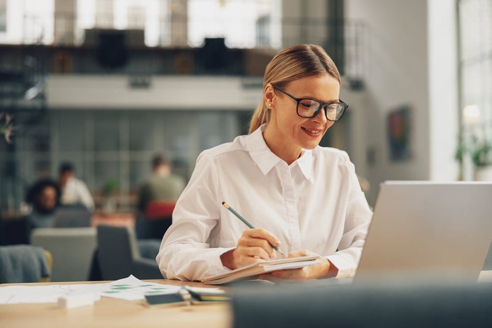 Womanofficeworkerworkingonlaptopandmakingnoteswhile